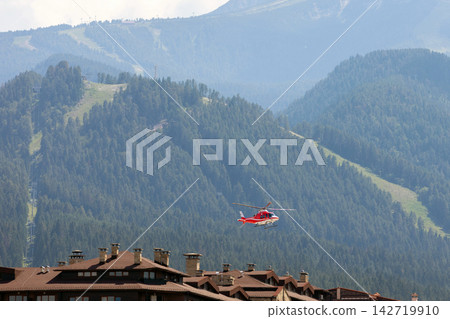 Bansko, Bulgaria, red emergency medical service helicopter flying over town, green Pirin mountains Bansko, Bulgaria, red emergency medical service helicopter flying over town, green Pirin mountains 142719910