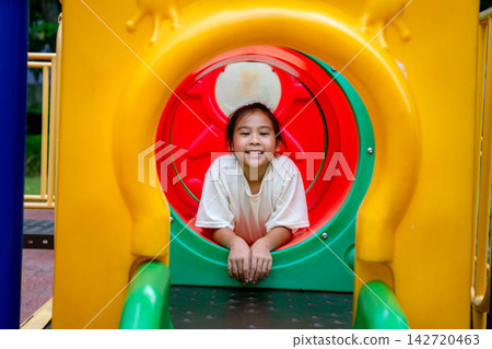 Happy Asian girl smiling and peeking out of round tube slide on playground 142720463