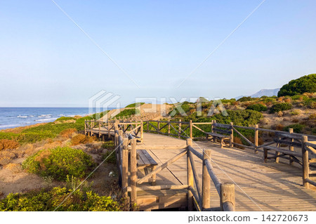 Wooden walkway leads through protected sand dunes covered with native coastal vegetation toward the Mediterranean shoreline in Cabopino, Spain Wooden walkway leads through protected sand dunes covered with native coastal vegetation toward the Mediterranean shoreline in Cabopino, Spain 142720673