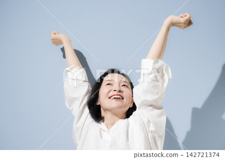 A woman in her 50s stretches with both arms raised high against a light blue background, smiling and enjoying a healthy lifestyle and refreshment. A woman in her 50s stretches with both arms raised high against a light blue background, smiling and enjoying a healthy lifestyle and refreshment. 142721374