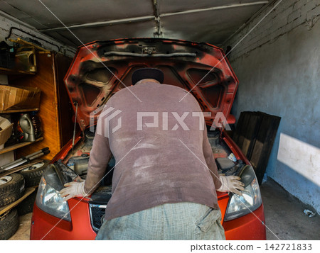 Man in work gloves and dirty clothes leaning over a car with its hood open, checking the engine during an auto service in a messy private garage. 142721833
