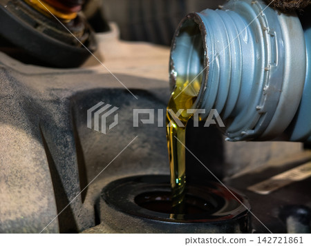 Golden liquid engine oil pouring from a grey plastic bottle neck into a car engine's oil filler opening, signifying routine automotive maintenance and lubrication for vehicle longevity. Golden liquid engine oil pouring from a grey plastic bottle neck into a car engine's oil filler opening, signifying routine automotive maintenance and lubrication for vehicle longevity. 142721861
