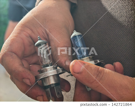 Mechanic hands holding and comparing two automotive halogen headlight bulbs, a clear old one and a new blue-tinted one, demonstrating car maintenance and replacement. 142721864