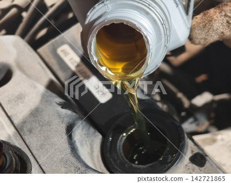 Mechanic pouring golden motor oil from a bottle into the car engine's filler neck, performing a crucial automotive service for vehicle longevity and optimal performance. Mechanic pouring golden motor oil from a bottle into the car engine's filler neck, performing a crucial automotive service for vehicle longevity and optimal performance. 142721865