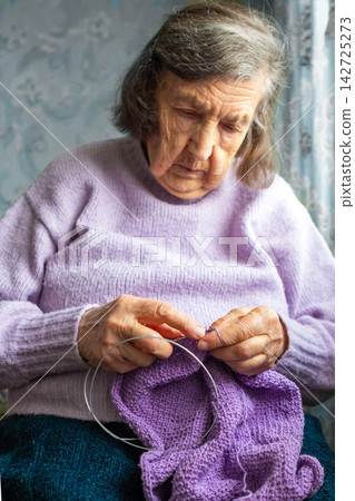 Elderly woman knitting a lavender scarf at home, focused senior enjoying a quiet hobby Elderly woman knitting a lavender scarf at home, focused senior enjoying a quiet hobby 142725273