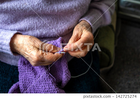 Elderly hands knitting purple yarn by a window, calm hobby and handmade comfort Elderly hands knitting purple yarn by a window, calm hobby and handmade comfort 142725274