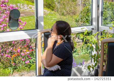 Thoughtful woman sitting in greenhouse looking through windows at blooming garden Thoughtful woman sitting in greenhouse looking through windows at blooming garden 142725351
