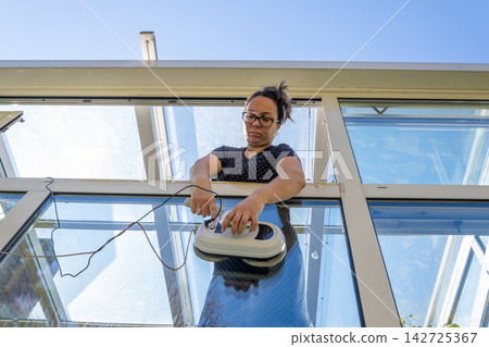 Focused woman operating a robotic window cleaner from a glass balcony Focused woman operating a robotic window cleaner from a glass balcony 142725367