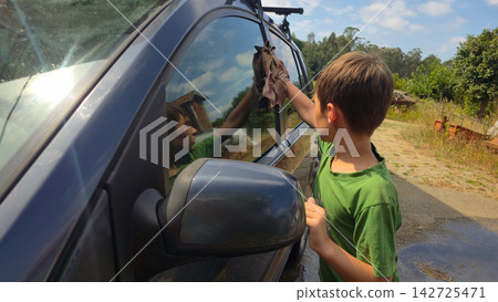 Child cleans a family car window during an outdoor home car wash. Concepts: household chores, teaching responsibility, family teamwork, practical life skills, vehicle care. Child cleans a family car window during an outdoor home car wash. Concepts: household chores, teaching responsibility, family teamwork, practical life skills, vehicle care. 142725471