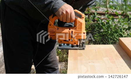 Sawing a wooden beam with an orange electric jigsaw. A man holds the tool while performing carpentry work outdoors. Close-up of hands, flying sawdust, and wood texture. Sawing a wooden beam with an orange electric jigsaw. A man holds the tool while performing carpentry work outdoors. Close-up of hands, flying sawdust, and wood texture. 142726147