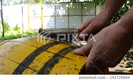 Cleaning an old wooden barrel with a wire brush. A man's hands carefully scrape dirt and rust off the yellow staves and black hoops in the yard. A close-up of the cooperage restoration process. Cleaning an old wooden barrel with a wire brush. A man's hands carefully scrape dirt and rust off the yellow staves and black hoops in the yard. A close-up of the cooperage restoration process. 142726604