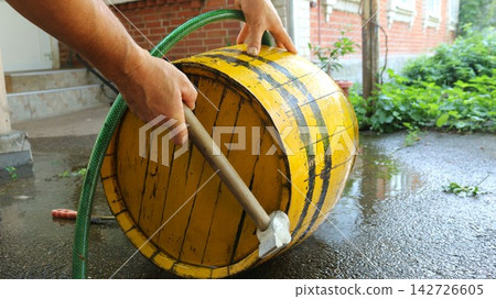 Preparing a wooden barrel for filling with distillate. A man's hands drive metal hoops down with a hammer on the wet asphalt of a courtyard. A green garden hose lies nearby Preparing a wooden barrel for filling with distillate. A man's hands drive metal hoops down with a hammer on the wet asphalt of a courtyard. A green garden hose lies nearby 142726605