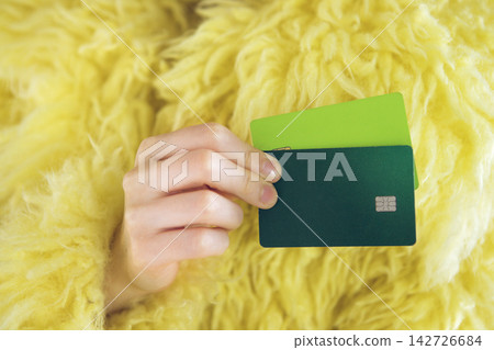 Hand holding two credit cards, one green and one dark blue, against a background of fluffy yellow fabric, emphasizing modern financial transactions and consumerism Hand holding two credit cards, one green and one dark blue, against a background of fluffy yellow fabric, emphasizing modern financial transactions and consumerism 142726684