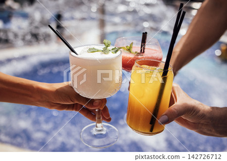 Three colorful cocktails held by hands, featuring a white frozen drink, a pink fruity beverage, and a yellow citrus drink, with a blurred pool background Three colorful cocktails held by hands, featuring a white frozen drink, a pink fruity beverage, and a yellow citrus drink, with a blurred pool background 142726712