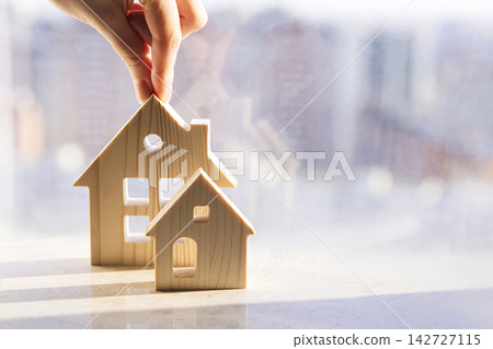 Hand placing wooden house models on a marble surface with a blurred cityscape visible through a window in the background, representing real estate and home concepts Hand placing wooden house models on a marble surface with a blurred cityscape visible through a window in the background, representing real estate and home concepts 142727115