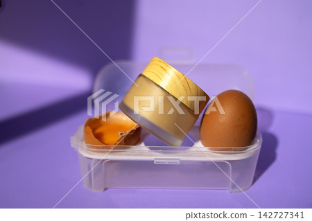 Brown egg placed beside a wooden jar and eggshell inside a transparent container on a purple background with soft lighting creating shadows Brown egg placed beside a wooden jar and eggshell inside a transparent container on a purple background with soft lighting creating shadows 142727341