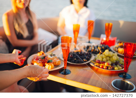 Group of friends enjoying a summer gathering on a boat with colorful drinks and a variety of fruits and pastries on a wooden table by the water Group of friends enjoying a summer gathering on a boat with colorful drinks and a variety of fruits and pastries on a wooden table by the water 142727370