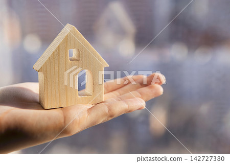 Wooden house model held in a hand against a blurred cityscape background, symbolizing real estate investment and home ownership concepts 142727380
