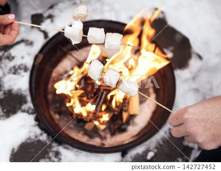 Two hands holding skewers with marshmallows roasting over a fire pit surrounded by snow, creating a cozy outdoor winter scene Two hands holding skewers with marshmallows roasting over a fire pit surrounded by snow, creating a cozy outdoor winter scene 142727452