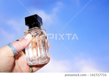 Hand holding a clear glass perfume bottle with a black cap against a bright blue sky, showcasing the elegant design and transparency of the fragrance container Hand holding a clear glass perfume bottle with a black cap against a bright blue sky, showcasing the elegant design and transparency of the fragrance container 142727710