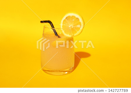 Refreshing lemonade drink in a clear glass with a lemon slice and black straw on a vibrant yellow background, perfect for summer beverage ideas and recipes 142727789