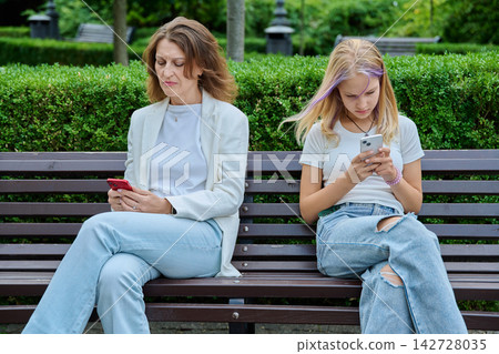 Mother and daughter sitting on bench ignored using smartphones outdoors Mother and daughter sitting on bench ignored using smartphones outdoors 142728035