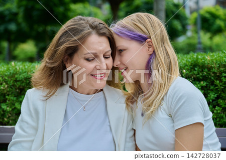 Teenage daughter whispering secret to smiling mature mother sitting outdoors in park Teenage daughter whispering secret to smiling mature mother sitting outdoors in park 142728037