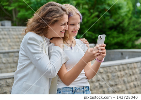 Mother leaning on teenage daughter shoulder looking at smartphone outdoors Mother leaning on teenage daughter shoulder looking at smartphone outdoors 142728040