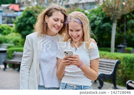 Happy mother and teenage daughter looking at smartphone together outdoors in city park Happy mother and teenage daughter looking at smartphone together outdoors in city park 142728046