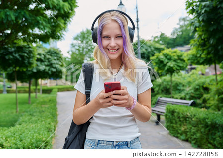 Smiling teenage schoolgirl using smartphone with headphones outdoors in park 142728058