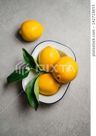Fresh lemons with green leaves in white enamel dish on textured gray background, natural citrus still life Fresh lemons with green leaves in white enamel dish on textured gray background, natural citrus still life 142728655