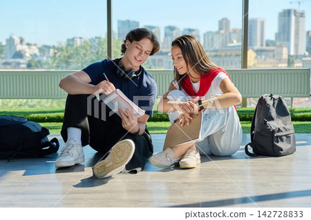 Two young students studying together with notebooks sitting on floor near big window Two young students studying together with notebooks sitting on floor near big window 142728833