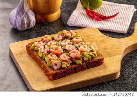 Shrimp and avocado toast on wooden cutting board with garlic and chili peppers in background Shrimp and avocado toast on wooden cutting board with garlic and chili peppers in background 142730748