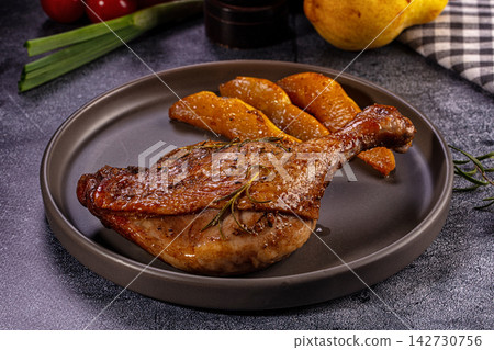 Roasted duck leg with rosemary and caramelized pear slices on dark ceramic plate, green onion and checkered napkin in background 142730756