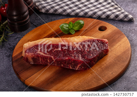 Raw marbled beef steak on round wooden cutting board with fresh basil leaves, black pepper mill, and checkered cloth background Raw marbled beef steak on round wooden cutting board with fresh basil leaves, black pepper mill, and checkered cloth background 142730757