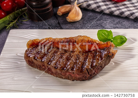Grilled steak with charred crust and fresh basil garnish on white textured plate, garlic and cherry tomatoes in background 142730758