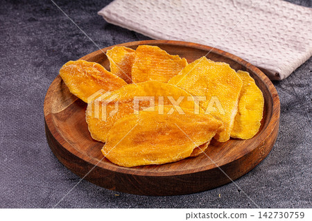 Dried mango slices on round wooden plate with textured cloth background, warm golden tones and rustic presentation Dried mango slices on round wooden plate with textured cloth background, warm golden tones and rustic presentation 142730759