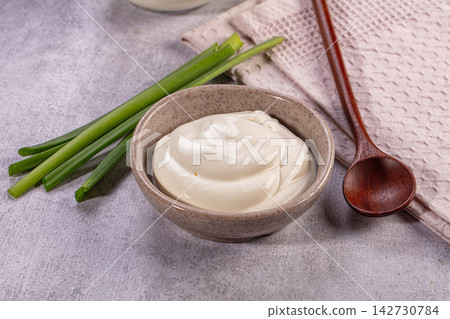Creamy white sauce in speckled ceramic bowl with fresh green onions and wooden spoon on textured gray surface Creamy white sauce in speckled ceramic bowl with fresh green onions and wooden spoon on textured gray surface 142730784