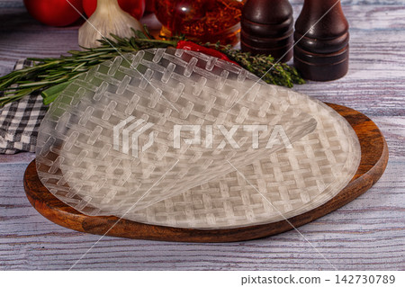 Transparent rice paper sheets with woven texture on wooden board, surrounded by fresh rosemary, garlic, tomatoes, and spice jars Transparent rice paper sheets with woven texture on wooden board, surrounded by fresh rosemary, garlic, tomatoes, and spice jars 142730789