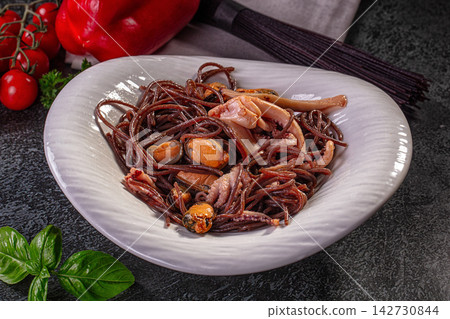 Dark squid ink spaghetti with squid rings and mussels served on white plate, fresh basil and cherry tomatoes on dark stone background Dark squid ink spaghetti with squid rings and mussels served on white plate, fresh basil and cherry tomatoes on dark stone background 142730844