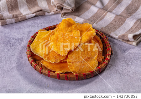 Dried mango slices in woven bamboo basket with red trim, set on gray surface with striped fabric background, warm golden tones and soft lighting Dried mango slices in woven bamboo basket with red trim, set on gray surface with striped fabric background, warm golden tones and soft lighting 142730852