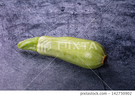 Fresh green zucchini with intact stem on dark textured surface, natural lighting highlights skin texture and subtle color variations Fresh green zucchini with intact stem on dark textured surface, natural lighting highlights skin texture and subtle color variations 142730902