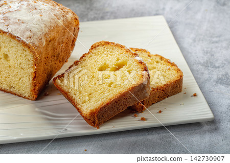 Sliced pound cake with golden crust and soft crumb on white plate, dusted with powdered sugar against gray textured background Sliced pound cake with golden crust and soft crumb on white plate, dusted with powdered sugar against gray textured background 142730907