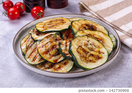 Grilled zucchini slices with char marks and spice seasoning on white ceramic plate, cherry tomatoes and striped napkin in background Grilled zucchini slices with char marks and spice seasoning on white ceramic plate, cherry tomatoes and striped napkin in background 142730908