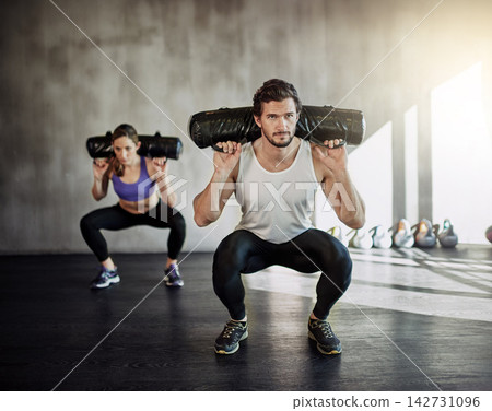 Man, woman and squat with sandbag for gym exercise or leg strength for muscle building, discipline or athlete. Personal trainer, client and weightlifting class for sports goals, workout or fitness Man, woman and squat with sandbag for gym exercise or leg strength for muscle building, discipline or athlete. Personal trainer, client and weightlifting class for sports goals, workout or fitness 142731096
