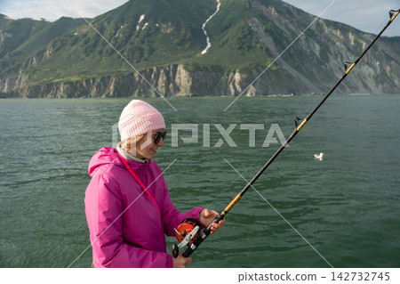 Female traveler fishes from a boat during a scenic sea journey in Kamchatka with rugged green coast behind. Fishing adventure, active tourism, outdoor leisure, exploration and nature travel. Female traveler fishes from a boat during a scenic sea journey in Kamchatka with rugged green coast behind. Fishing adventure, active tourism, outdoor leisure, exploration and nature travel. 142732745