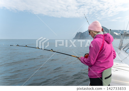 Female angler fishes from a boat beside rugged Kamchatka coast. Scenic sea trip, marine travel, recreational fishing, outdoor leisure, active journey, nature tourism and adventure travel. Female angler fishes from a boat beside rugged Kamchatka coast. Scenic sea trip, marine travel, recreational fishing, outdoor leisure, active journey, nature tourism and adventure travel. 142733833