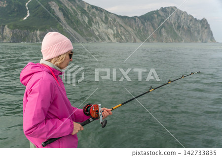 Woman fishes against dramatic mountain coast during a sea trip in Kamchatka. Marine journey, recreational fishing, active travel, outdoor recreation, adventure tourism and nature experience. 142733835