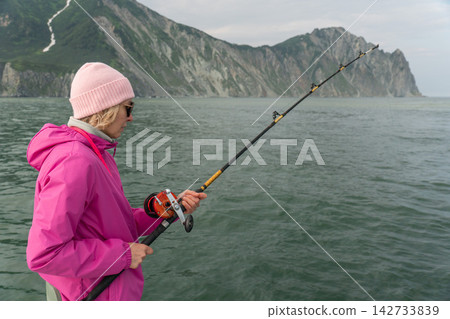 Female traveler fishes from a boat during a scenic sea trip in Kamchatka. Recreational fishing, marine travel, outdoor leisure, adventure tourism, active journey and coastal exploration. Female traveler fishes from a boat during a scenic sea trip in Kamchatka. Recreational fishing, marine travel, outdoor leisure, adventure tourism, active journey and coastal exploration. 142733839