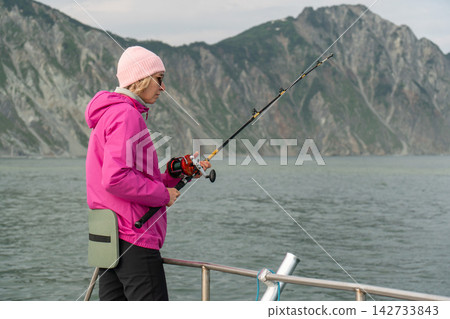 Woman enjoys fishing from a boat along rugged Kamchatka coast. Sea trip, recreational fishing, active travel, outdoor leisure, marine adventure, nature tourism and memorable journey. Woman enjoys fishing from a boat along rugged Kamchatka coast. Sea trip, recreational fishing, active travel, outdoor leisure, marine adventure, nature tourism and memorable journey. 142733843